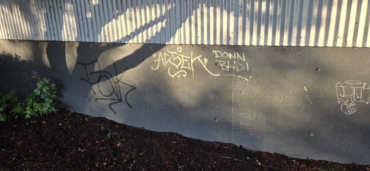 Real spray-painted graffiti tags and a black bubble-style tag on a concrete retaining wall near a home in McMinnville, Oregon — actual property vandalism a homeowner has to clean up